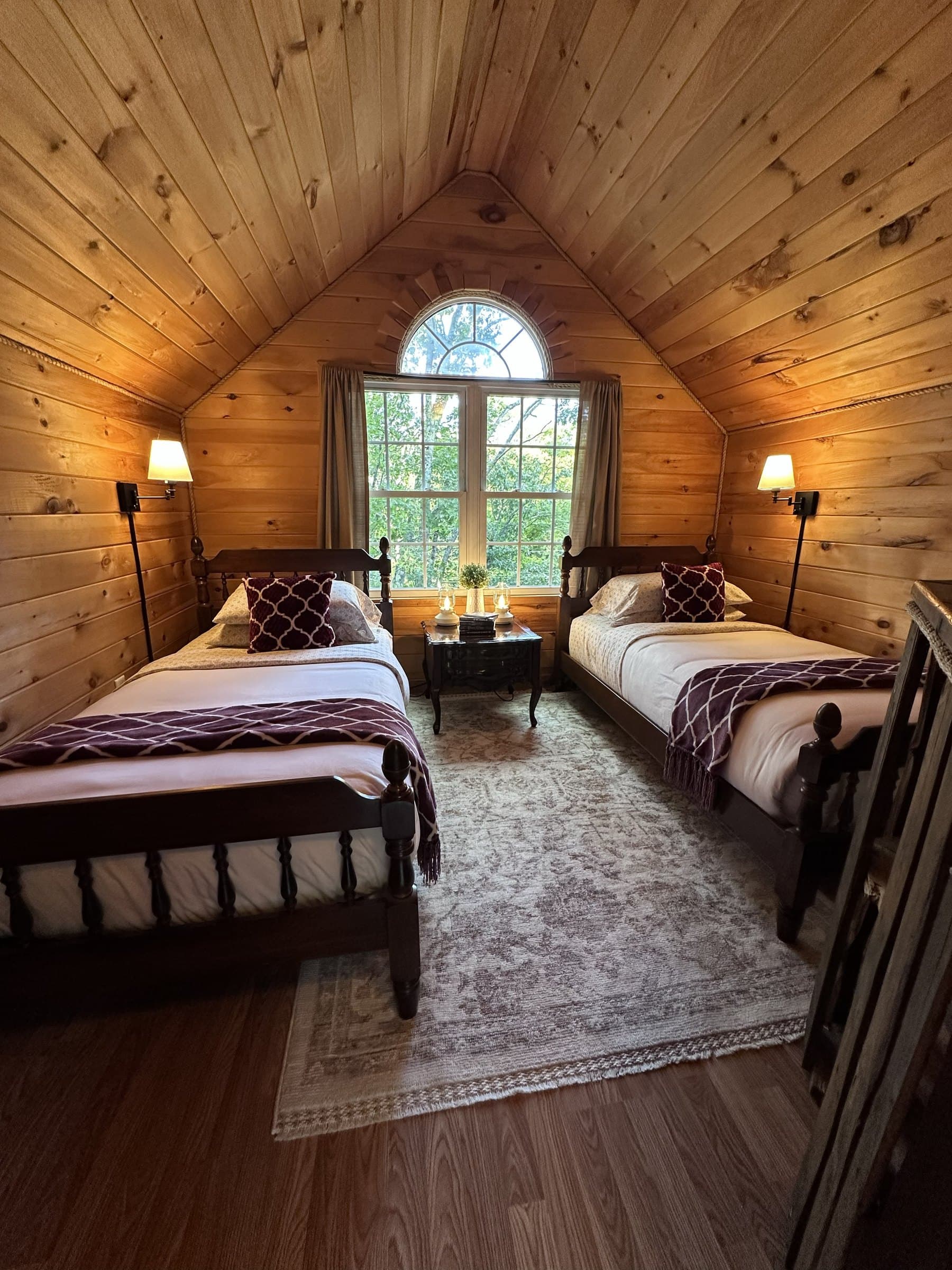 Two twin beds with plum throws and lit wall lamps under the loft's arched window