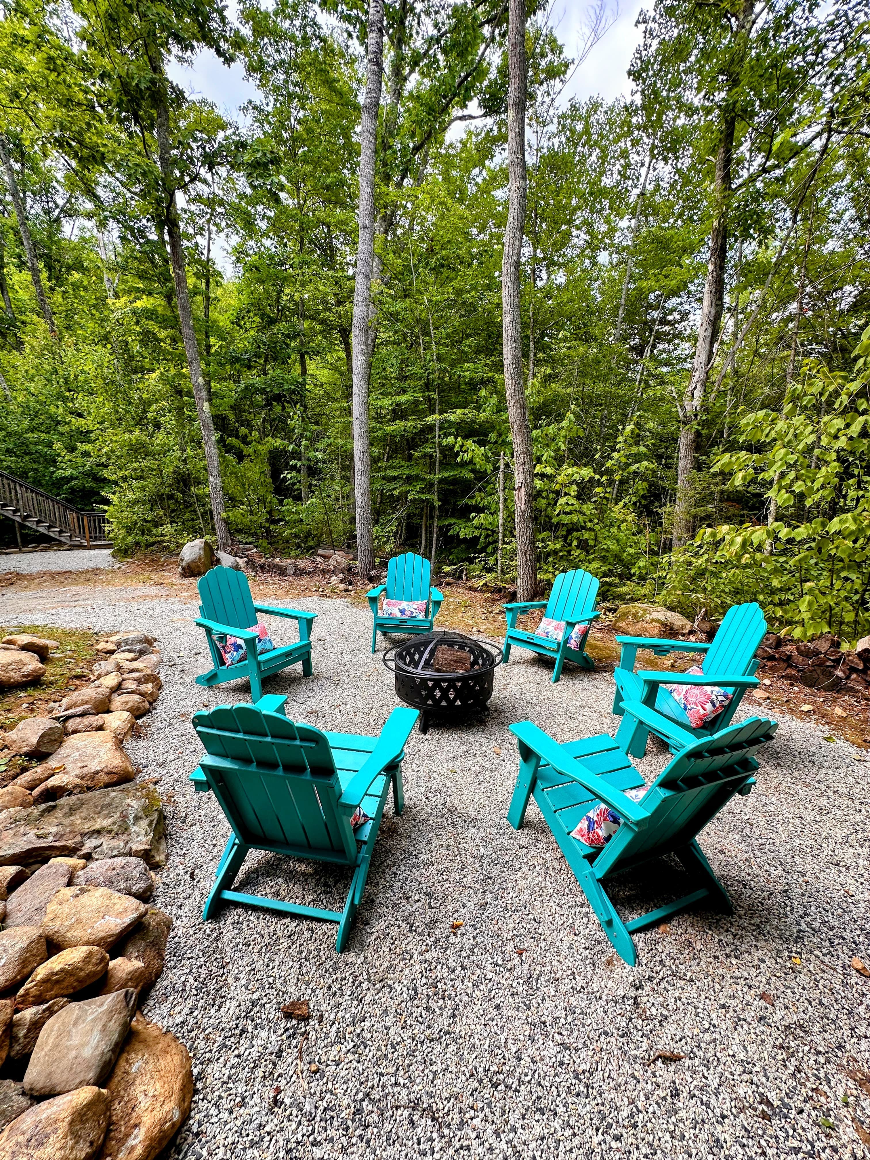 Teal Adirondack chairs around a firepit on a gravel pad among the trees